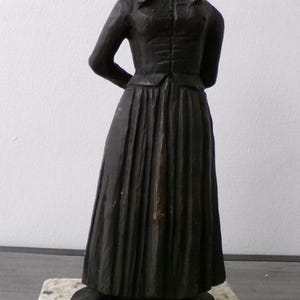 Könnte beinhalten: Eine dunkelbraune, monochrome Skulptur einer stehenden Figur, die einen langen, plissierten Rock, eine taillierte Jacke und eine Kopfbedeckung trägt. Die Figur ist auf einem rechteckigen, gesprenkelten beige-braunen Steinsockel montiert.