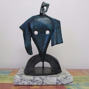 May include: A dark blue-green abstract metal sculpture featuring a central head-like form with two circular cutouts for eyes, mounted on a rectangular speckled gray and white granite base. The sculpture has a smooth, weathered metallic finish.