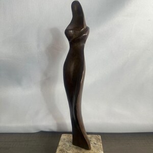 May include: A tall, slender, dark brown wooden sculpture featuring a smooth, abstract, elongated form that curves gracefully. The sculpture is mounted on a square, light-colored, speckled stone base with a textured, natural appearance.