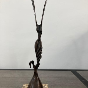 May include: A tall, dark bronze-toned sculpture featuring an elongated, abstract form with two upward-reaching, wing-like extensions. The sculpture has a smooth, metallic finish and is mounted on a rectangular, speckled beige and brown stone base.