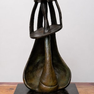 May include: A bronze sculpture featuring a dark, textured, abstract form with a hollow, pear-shaped base and an upper section composed of vertical bars supporting a small, T-shaped top piece, all mounted on a rectangular, polished dark stone base.