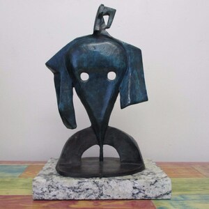 May include: A dark blue-green abstract metal sculpture featuring a central head-like form with two circular cutouts for eyes, mounted on a rectangular speckled gray and white granite base. The sculpture has a smooth, weathered metallic finish.