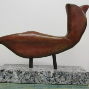 May include: A smooth, reddish-brown abstract sculpture shaped like a bird, mounted on two thin dark metal rods. The sculpture is attached to a rectangular base made of speckled gray and white granite with a polished, reflective surface finish.