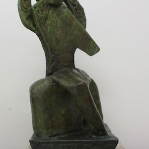 May include: A bronze-toned sculpture with a textured, weathered surface featuring an abstract, elongated form that rises from a rectangular base. The sculpture is mounted on a thick, speckled, light-colored stone plinth with a polished finish.