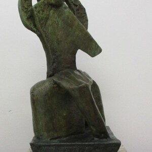 May include: A bronze-toned sculpture with a textured, weathered surface featuring an abstract, elongated form that rises from a rectangular base. The sculpture is mounted on a thick, speckled, light-colored stone plinth with a polished finish.