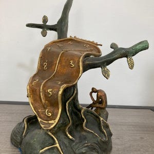 Puede incluir: Una escultura en tonos bronce que presenta un reloj derretido sobre una rama de árbol oscura y texturizada. Una pequeña figura sentada descansa en la base del árbol. La esfera muestra números dorados en relieve, y la pieza se asienta sobre una base oscura e irregular con detalles en oro metálico en los bordes.