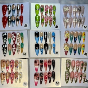 May include: Nine sets of medium-length almond-shaped press-on nails displayed on white cards. Designs include red, chrome, floral, butterfly, bow, polka dot, cross, star, and gemstone patterns in various colors like green, blue, pink, and black.