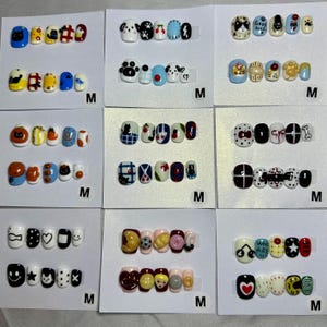 May include: Nine sets of press-on nails displayed on white cards, each labeled with an M. The nails feature diverse colorful designs including cats, food items, geometric patterns, hearts, stars, and text like Good Luck and Who Are You.