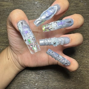 May include: Long square-shaped artificial nails featuring a light blue and silver marbled base. The nails are decorated with various embellishments including small silver studs, iridescent sequins, tiny seashell charms, and a small white flower.