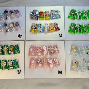 Op de afbeelding: Zes sets kunstnagels gepresenteerd op witte kaarten, elk gemarkeerd met een M. De ontwerpen bevatten diverse kleuren waaronder transparant, geel, groen en roze, versierd met 3D-bedels zoals sterren, harten, schedels, sleutels en kleine dierfiguren.