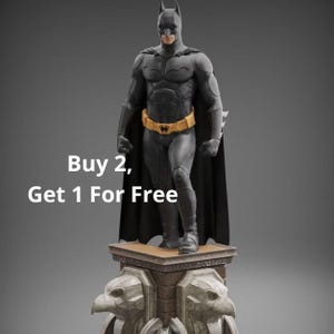 May include: A Batman action figure standing on a stone-textured pedestal with eagle carvings. The figure wears a black suit with a yellow utility belt and a long cape. The text Buy 2, Get 1 For Free is displayed on the left side of the image.