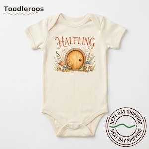 May include: A cream-colored short-sleeve baby bodysuit featuring a graphic print of a round wooden door surrounded by mushrooms and foliage. The text HALFLING is printed in a brown serif font above the door illustration on the front center.