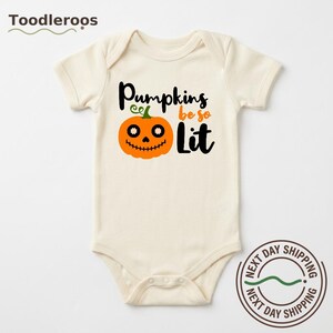 Może przedstawiać: Kremowe body niemowlęce z krótkim rękawem z grafiką pomarańczowej dyni z czarnymi oczami i zszytymi ustami. Napis Pumpkins be so Lit jest nadrukowany czarnymi i pomarańczowymi literami, łączącymi pismo odręczne z drukowanym na przodzie.