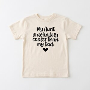 Puede incluir: Una camiseta de manga corta de color crema con el texto en negro My Aunt is definitely cooler than my Dad, centrado sobre un pequeño icono de corazón negro. La prenda tiene cuello redondo y un corte estándar de tela lisa.