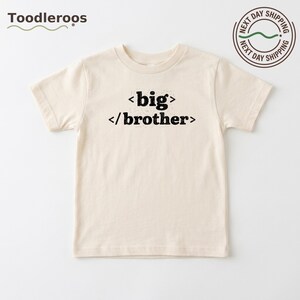 May include: A cream-colored short-sleeve t-shirt featuring the black text big brother enclosed in coding-style angle brackets. The design is centered on the chest of the shirt, which has a crew neckline and a standard straight hemline.