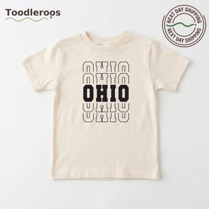 May include: A cream-colored short-sleeve t-shirt featuring the word OHIO printed in a stacked, repeating design. The central OHIO is in solid black, while the four surrounding instances of the word are outlined in black, creating a layered effect.
