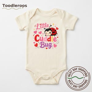 May include: A cream-colored short-sleeve baby bodysuit featuring a graphic of a red and black ladybug with a winking expression. The text Little Cuddle Bug is printed in colorful, decorative lettering surrounded by small pink and red hearts.