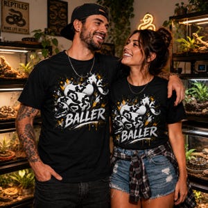 May include: A black t-shirt featuring a graphic of a coiled snake with white and black scales, surrounded by yellow and white paint splatter effects, with the word BALLER printed in a bold, distressed white font across the centre of the design.
