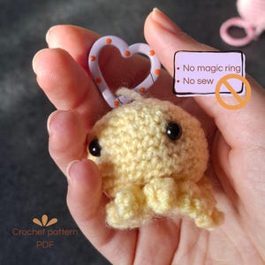 Op de afbeelding: Een lichtgele gehaakte octopus sleutelhanger met zwarte plastic ogen bevestigd aan een lichtpaarse hartvormige karabijnhaak. Een tekstvak toont: No magic ring, No sew. De tekst Crochet pattern PDF is zichtbaar in de linkerbenedenhoek.
