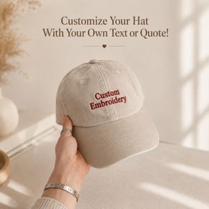 May include: A beige baseball cap featuring the embroidered text "Custom Embroidery" in dark red thread on the front panel. The text "Customize Your Hat With Your Own Text or Quote!" is printed above the cap in a dark brown serif font.