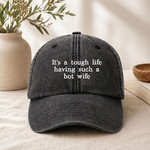 May include: A charcoal grey baseball cap with a distressed, washed-out finish. The front panel features white embroidered text that reads, "It's a tough life having such a hot wife." The cap has a curved brim and a structured crown with stitched seams.