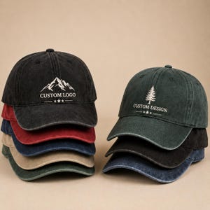 Op de afbeelding: Twee stapels honkbalpetten in diverse kleuren zoals zwart, rood, marineblauw, beige en donkergroen. De bovenste zwarte pet heeft een witte bergafbeelding en de tekst CUSTOM LOGO. De bovenste donkergroene pet toont een dennenboom en CUSTOM DESIGN.