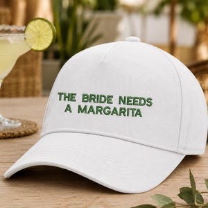 Może przedstawiać: Biała czapka z daszkiem z wyhaftowanym zielonym napisem THE BRIDE NEEDS A MARGARITA na przednim panelu. Czapka ma usztywnianą koronę, zakrzywiony daszek i guzik na górze, prezentując czysty design z wyraźnymi, ciemnozielonymi literami.