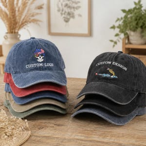 May include: A stack of five baseball caps in blue, red, navy, tan, and olive green, featuring an embroidered skull with an American flag hat and the text CUSTOM LOGO. Beside it, a stack of three black baseball caps with a fish graphic and CUSTOM DESIGN.