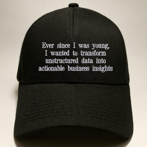 May include: A black baseball cap featuring white embroidered text that reads: Ever since I was young, I wanted to transform unstructured data into actionable business insights. The cap has a structured crown, a curved brim, and a top button.
