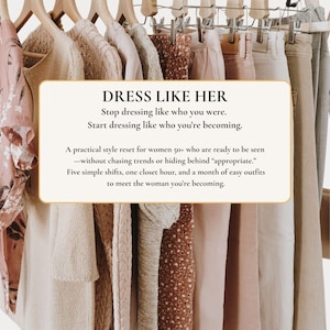 Op de afbeelding: Een kledingrek met diverse kledingstukken in neutrale tinten zoals beige, crème en zachtroze. Een rechthoekig bord op de voorgrond bevat de tekst: DRESS LIKE HER. Stop dressing like who you were. Start dressing like who you're becoming.