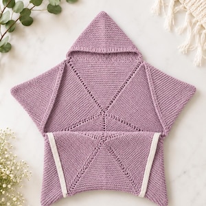 May include: A star-shaped knitted blanket in a dusty mauve colour featuring a pointed hood at the top. The garment is constructed with a garter stitch texture and includes white trim along the two bottom points, creating a wrap-style design.