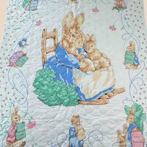 May include: A white quilted blanket featuring illustrations of Peter Rabbit characters. The center shows a rabbit in a blue dress holding two smaller rabbits. Surrounding characters include Tiggy Winkle, Jemima Puddle Duck, Benjamin Bunny, Tom Kitten, and Flopsy Bunnies, each holding a book with their name printed on the cover.
