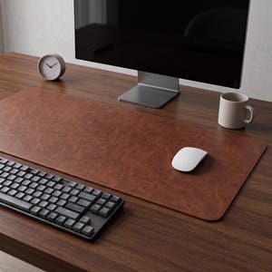 Peut inclure: Un sous-main rectangulaire en cuir marron à la surface texturée posé sur un bureau en bois. Le tapis se trouve sous un écran d'ordinateur noir, un clavier mécanique gris foncé, une souris blanche, un stylo argenté, une petite horloge et une tasse.