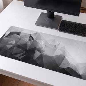 May include: A rectangular desk mat featuring a monochromatic geometric pattern of triangles in varying shades of black, grey, and white. The design transitions from dark tones on the left side to lighter tones on the right side across the surface.