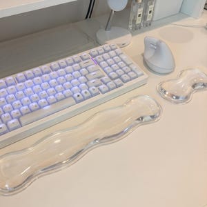 May include: A white mechanical keyboard with backlit keys, a white ergonomic vertical mouse, and two clear, wavy-shaped gel wrist rests. The setup includes a white magnetic charging stand and two small glass bottles on a white desk surface.