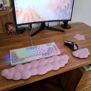Könnte beinhalten: Eine mechanische Tastatur mit weißen, rosa und violetten Tastenkappen und RGB-Hintergrundbeleuchtung, begleitet von einer langen, hellrosa wolkenförmigen Handballenauflage und einem kleineren passenden wolkenförmigen Mauspad auf einem Schreibtisch aus Holz.