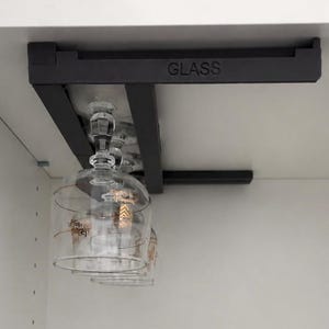 May include: A black metal under-cabinet rack with the word GLASS printed on the side, holding two clear glass stemware pieces. The glasses feature thin, horizontal gold-coloured lines and small gold-coloured graphic details around the bowl.