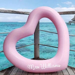 Puede incluir: Un flotador inflable grande en forma de corazón de color rosa claro con el texto "Mrs. Williams" impreso en letra cursiva blanca en la parte inferior frontal. El flotador está colocado sobre una cubierta de madera frente al mar azul.