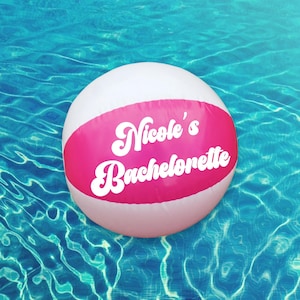 May include: A round, inflatable beach ball featuring a white base with a wide, horizontal hot pink band across the center. The text "Nicole's Bachelorette" is printed in a bold, white, retro-style cursive font across the pink section.