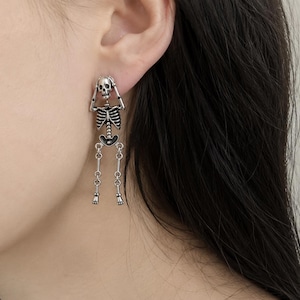 Peut inclure: Une boucle d'oreille pendante de couleur argentée représentant un squelette avec les mains posées sur la tête. Le squelette présente une cage thoracique et un crâne détaillés, avec des jambes articulées pendantes terminées par de petits pieds.