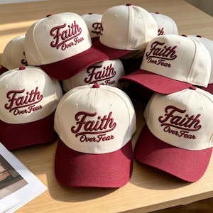 May include: A collection of baseball caps featuring a cream-colored crown and a dark red brim. Each cap displays the embroidered text Faith Over Fear in a dark red script font across the front panel, with a structured design and top button.
