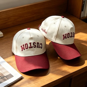 Könnte beinhalten: Zwei Baseballkappen mit cremeweißen Kronen und dunkelroten Schirmen. Auf der Vorderseite jeder Kappe ist das Wort CUSTOM in dunkelroten Großbuchstaben eingestickt. An der Seite der hinteren Kappe steht das Wort custom in roter Schreibschrift.