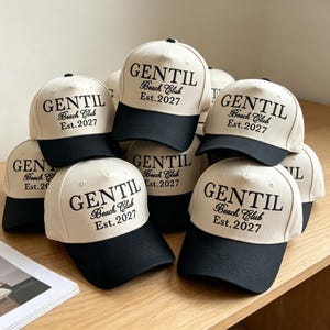 May include: A collection of baseball caps featuring a cream-colored crown and a black brim. Each cap displays the black embroidered text GENTIL Beach Club Est. 2027 on the front panel. The caps are arranged in a stacked formation on a flat surface.