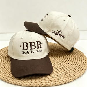 Pode incluir: Dois bonés de beisebol com copas em tom off-white e abas marrom-escuras. O boné da frente apresenta bordado marrom-escuro com o texto BBB e Body by becca. O boné de trás exibe a palavra custom em letra cursiva marrom-escura no painel lateral.