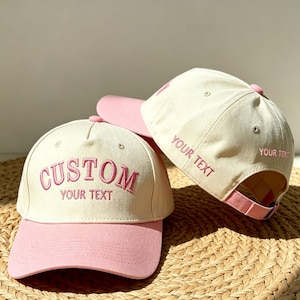 Op de afbeelding: Twee baseballpetten met een crèmekleurige kroon en een roze klep. De voorste pet toont de geborduurde roze tekst CUSTOM YOUR TEXT. De tweede pet, die erachter staat, heeft de geborduurde roze tekst YOUR TEXT op het zijpaneel.