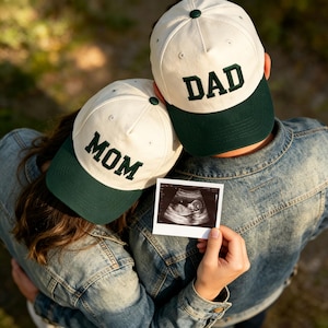 Może przedstawiać: Dwie czapki z daszkiem z kremowymi koronami i ciemnozielonymi daszkami. Czapka po lewej stronie ma wyhaftowane ciemnozielonymi literami słowo MOM, natomiast czapka po prawej stronie ma wyhaftowane ciemnozielonymi literami słowo DAD.