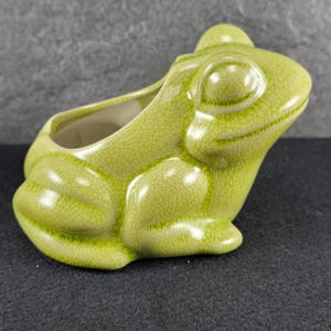 May include: A light green ceramic planter shaped like a frog with a glossy finish and a visible crackle glaze texture. The frog figure is depicted in a seated position with its head turned slightly to the side, featuring a hollow opening.