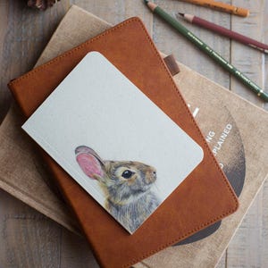 May include: A small white notebook featuring a detailed coloured pencil illustration of a brown rabbit with pink-tinted ears, resting on a tan leather-bound journal and a beige fabric-covered book, accompanied by three coloured pencils and pinecones.