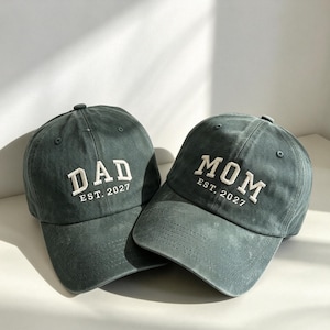Puede incluir: Dos gorras de béisbol de color verde oscuro con letras blancas bordadas. Una gorra muestra el texto DAD EST. 2027, mientras que la otra muestra el texto MOM EST. 2027. Ambas gorras tienen una corona estructurada, visera curva y ojales.