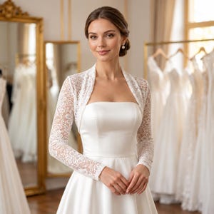 May include: A white lace bolero jacket with long sleeves and a floral pattern, worn over a strapless white satin dress. The bolero features a scalloped edge along the front opening and cuffs, providing a fitted silhouette over the bodice.